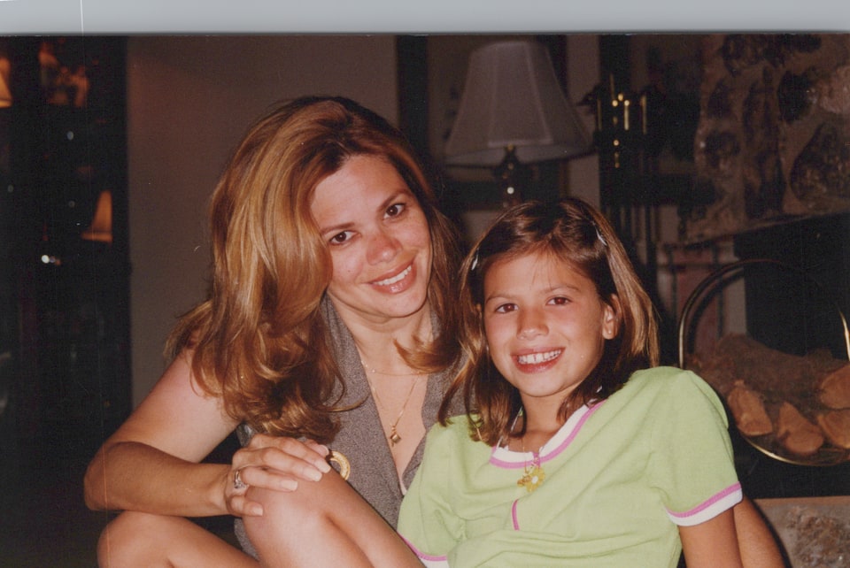 Xiomara smiles beside a young girl in a green shirt near a fireplace.