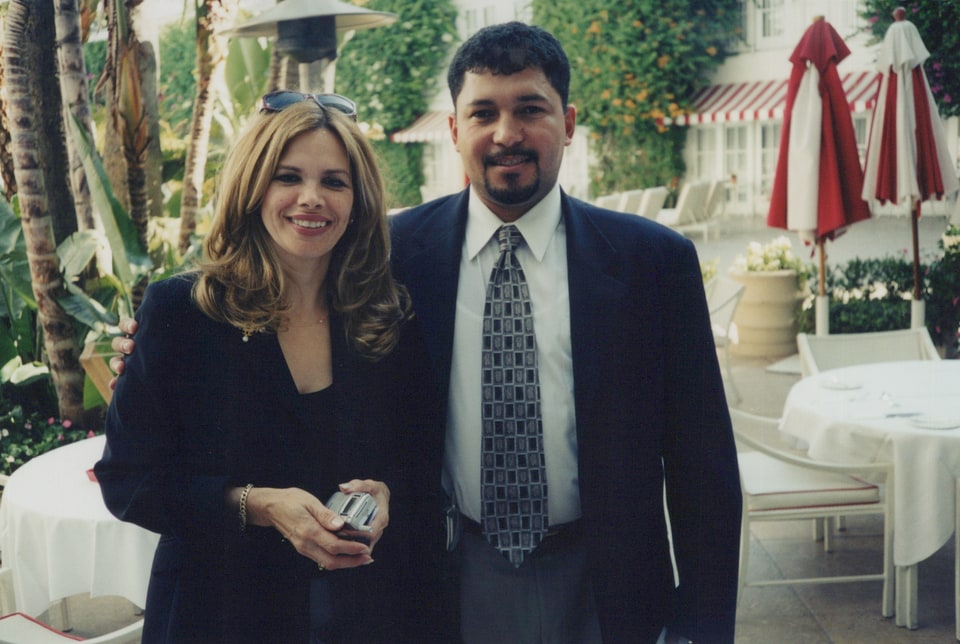 Xiomara and a man in suits smile outdoors beside red patio umbrellas.