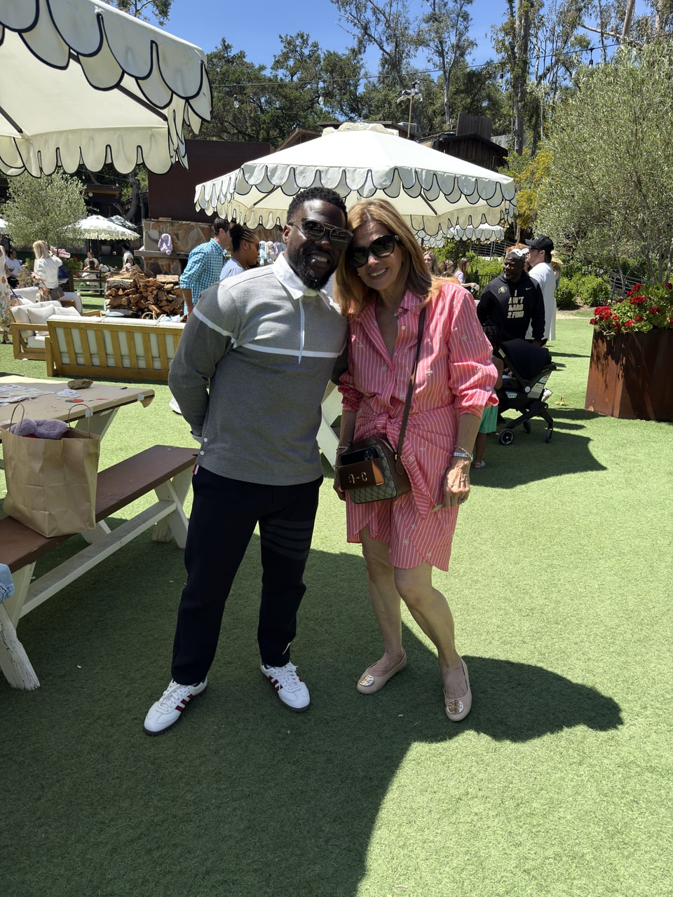 Xiomara in a pink striped dress and sunglasses smiles beside a man at an outdoor gathering.