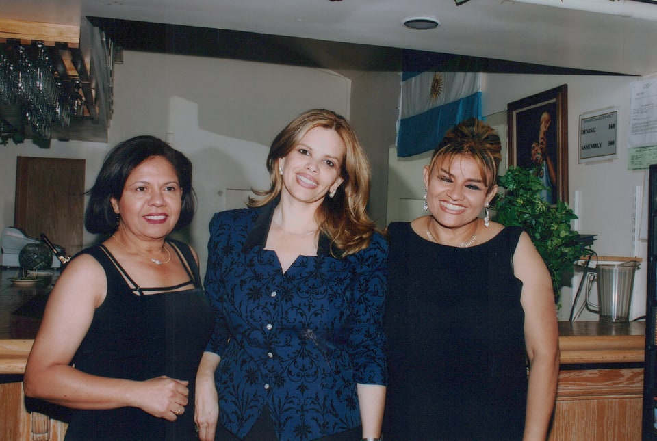 Xiomara in a blue patterned jacket stands between two women dressed in black.