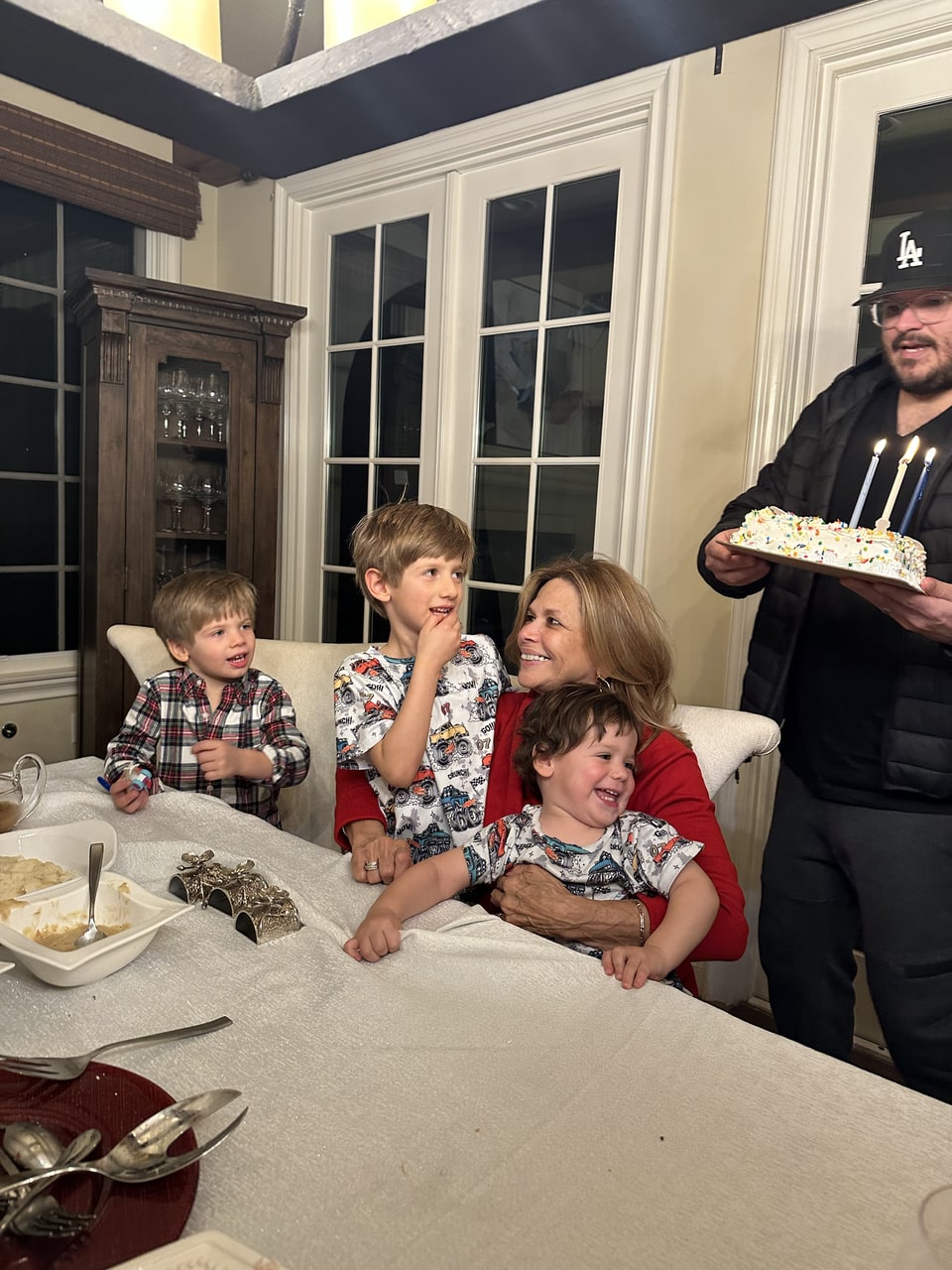 Xiomara in red smiles while holding a laughing child at a table with two other children and a person carrying a cake.