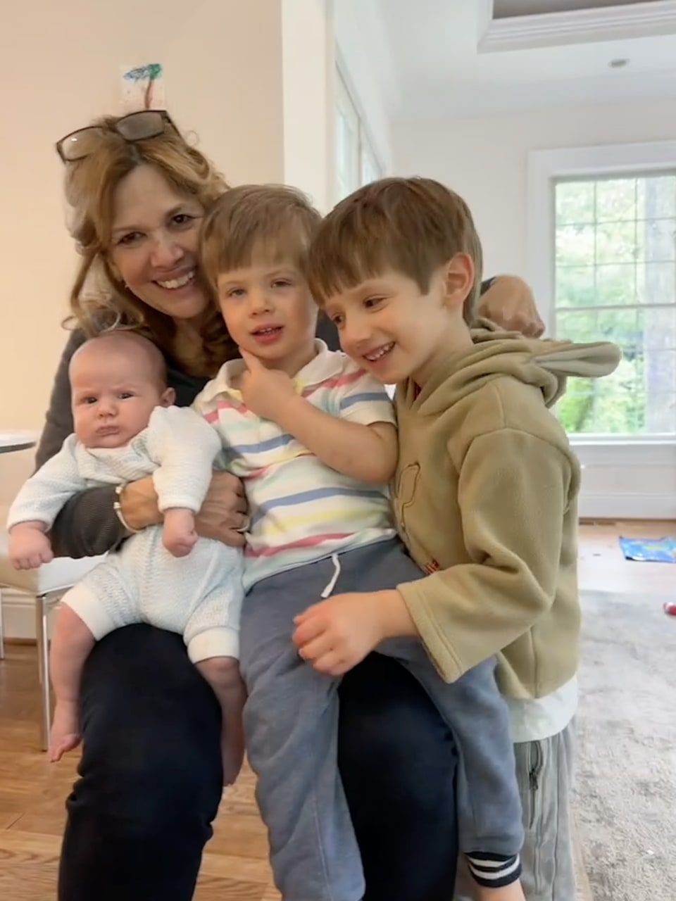 Xiomara smiles while holding a baby and sitting closely with two young boys indoors.
